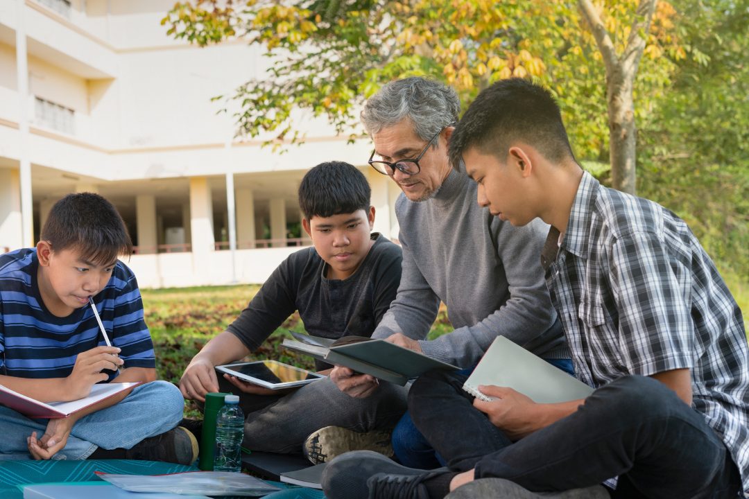 old teacher and group of teenager boys sitting together at school backyard,teacher holding a book explaining a lesson to the students, concept of education,